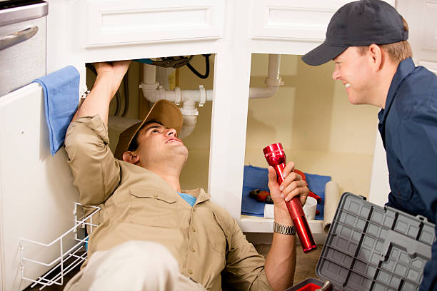 Plumbers, repairmen working under sink in home kitchen. Young adult, Latin and caucasian plumbers or repairmen working under a home kitchen sink. They have tools and a toolbox and wear uniforms. Water pipes, flashlight. plumbing team stock pictures, royalty-free photos & images