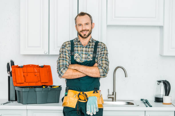 smiling handsome plumber standing with crossed arms and looking at camera in kitchen smiling handsome plumber standing with crossed arms and looking at camera in kitchen plumbing stock pictures, royalty-free photos & images
