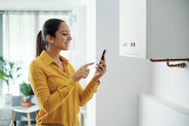 Woman managing her smart boiler using her phone Woman managing and programming her smart boiler using her smartphone, smart home concept water heater stock pictures, royalty-free photos & images