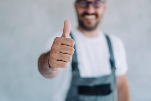 Young male worker in an overall uniform posing at his workplace. Shot of confident smiling professional handyman in overalls and white t-shirt standing in his workplace and showing thumb up. Focus is on the thumb. plumbing stock pictures, royalty-free photos & images