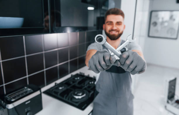 Young professional plumber in grey uniform standing on the kitchen and holding work equipment in hands